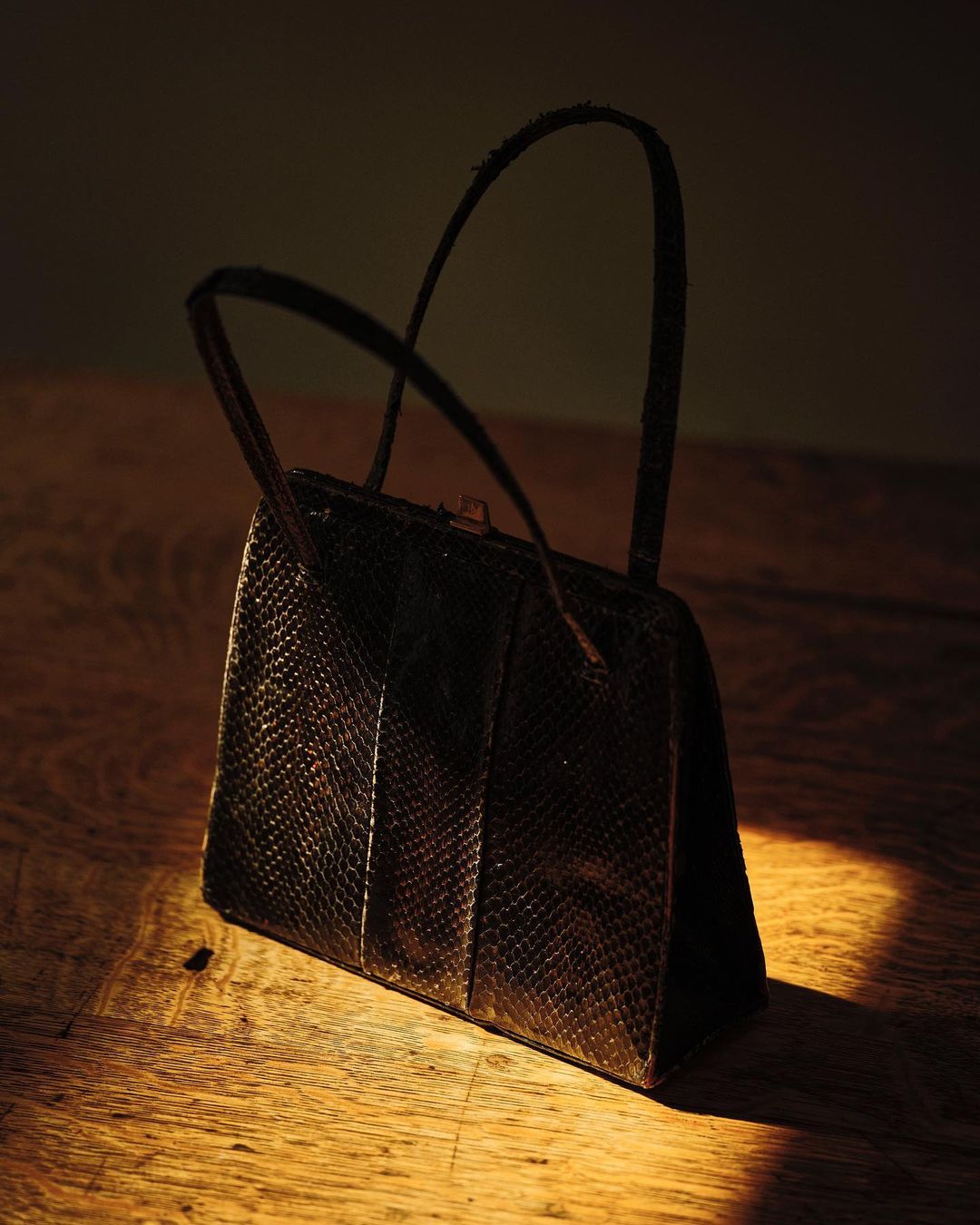 Reptile-patterned handbag with handles on wooden table in dark environment with warm, soft lighting creating highlights and shadows.