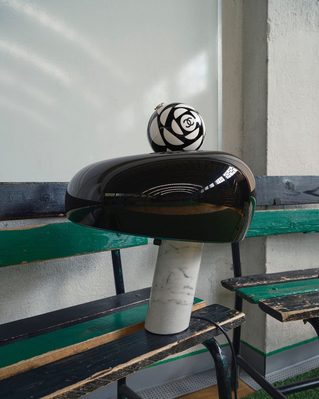 Chanel logo ball placed on top of a glossy black helmet-shaped object with a marble base, sitting on a weathered wood bench.