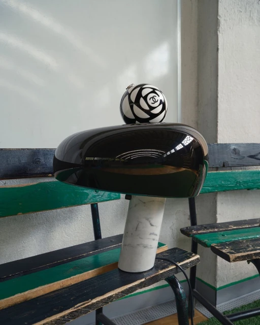 Chanel logo ball placed on top of a glossy black helmet-shaped object with a marble base, sitting on a weathered wood bench.