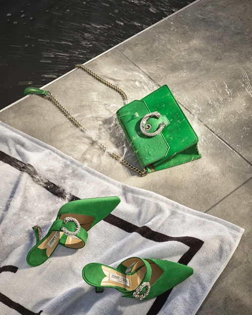 Overhead shot of a vibrant green Jimmy Choo purse and heels on a towel against tile, evoking high-fashion and luxury.