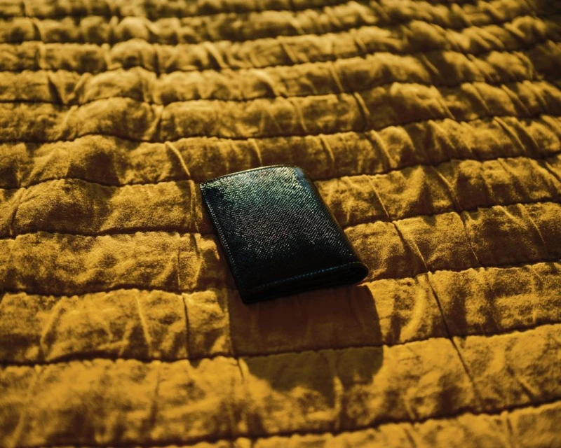 Close-up of a black wallet on textured yellow fabric. The wallet casts a shadow on the textured surface in warm, natural light.