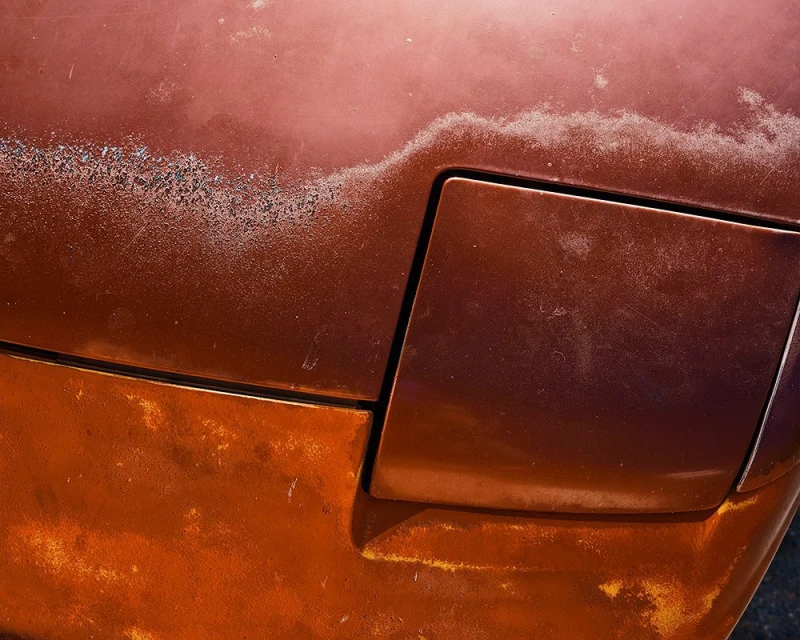 Close-up of the rusted hood and partially visible headlamp of a vintage car, with a brown-orange gradient and visible textures.