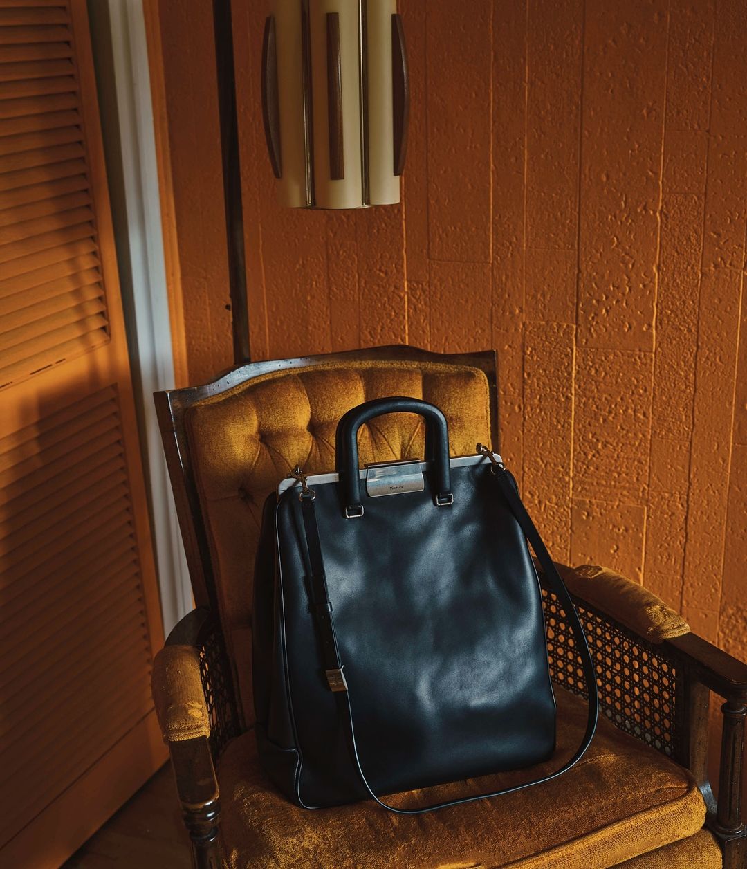 Black leather tote bag is displayed on a vintage upholstered chair in a room with orange walls and a modern light fixture.