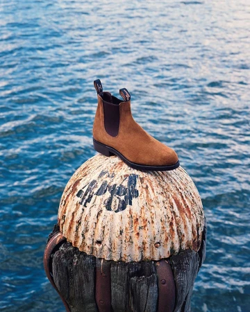 A single brown leather boot sits on a weathered bollard with the ocean in the background, creating a footwear fashion statement.