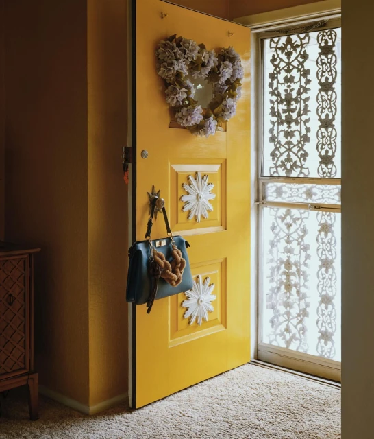 Bright yellow door with a floral heart wreath, purse, and keys hanging from it, with an ornate sheer curtain in the background.