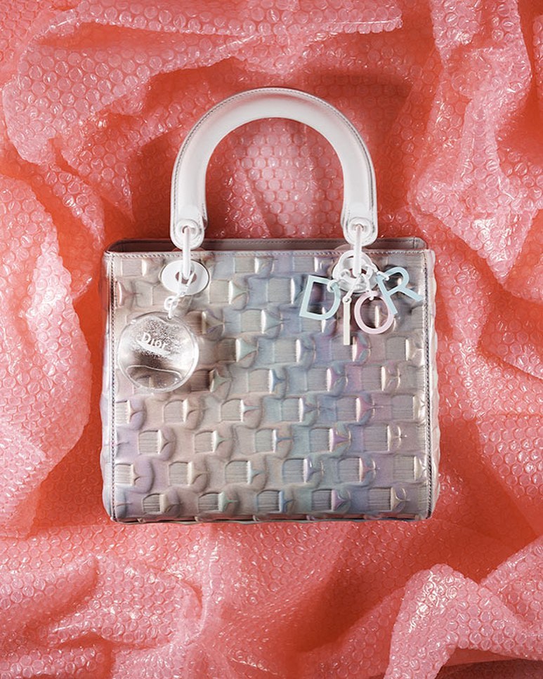Close-up of a translucent silver Lady Dior handbag with a white handle and silver Dior charms, placed on pink bubble wrap.