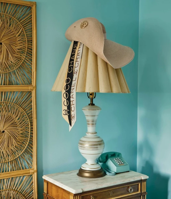 Interior still life with a straw hat and Chanel scarf draped over a lamp on a marble-topped wooden desk, set against a teal wall.