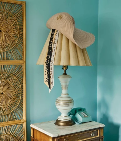 Interior still life with a straw hat and Chanel scarf draped over a lamp on a marble-topped wooden desk, set against a teal wall.