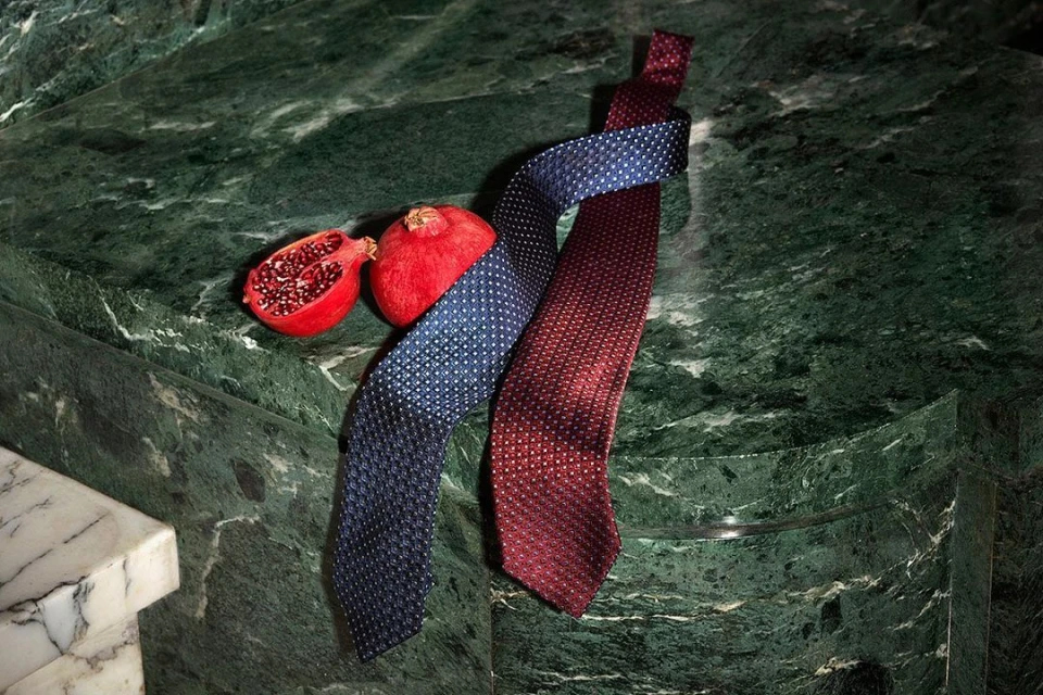 Still life composition featuring two patterned ties, one blue and one maroon, alongside fresh pomegranate fruit on a veined green surface.