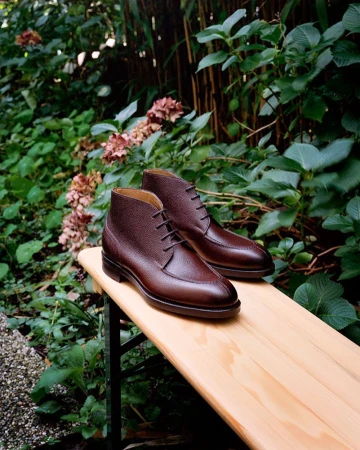 Elegant brown leather chukka boots displayed on a wooden bench outdoors, surrounded by lush green foliage and soft, natural lighting.