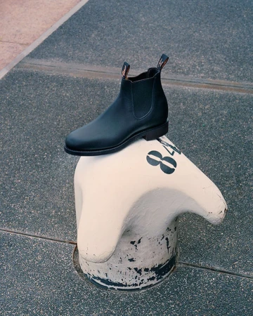 Black leather Chelsea boot on a white concrete bollard against a grey pavement backdrop, captured in natural lighting.