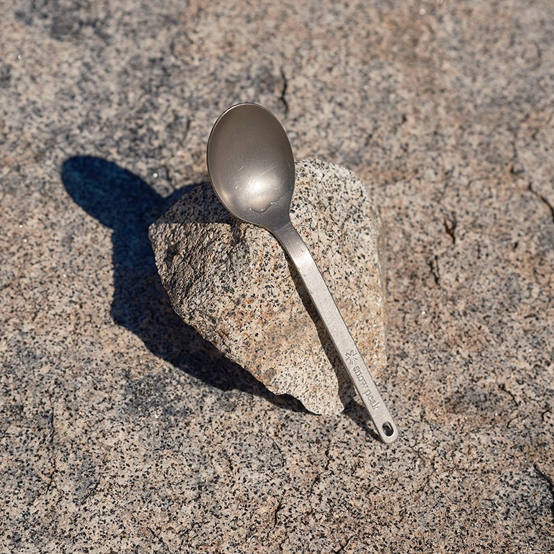 Titanium spoon resting on granite rock outdoors in bright sunlight casting a shadow. Features visible Snow Peak logo on handle.
