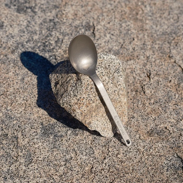 Titanium spoon resting on granite rock outdoors in bright sunlight casting a shadow. Features visible Snow Peak logo on handle.