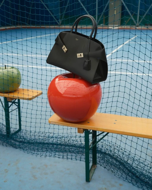 Studio shot of a black leather handbag on a red sphere, placed on a wooden bench against a blurred blue tennis court background.