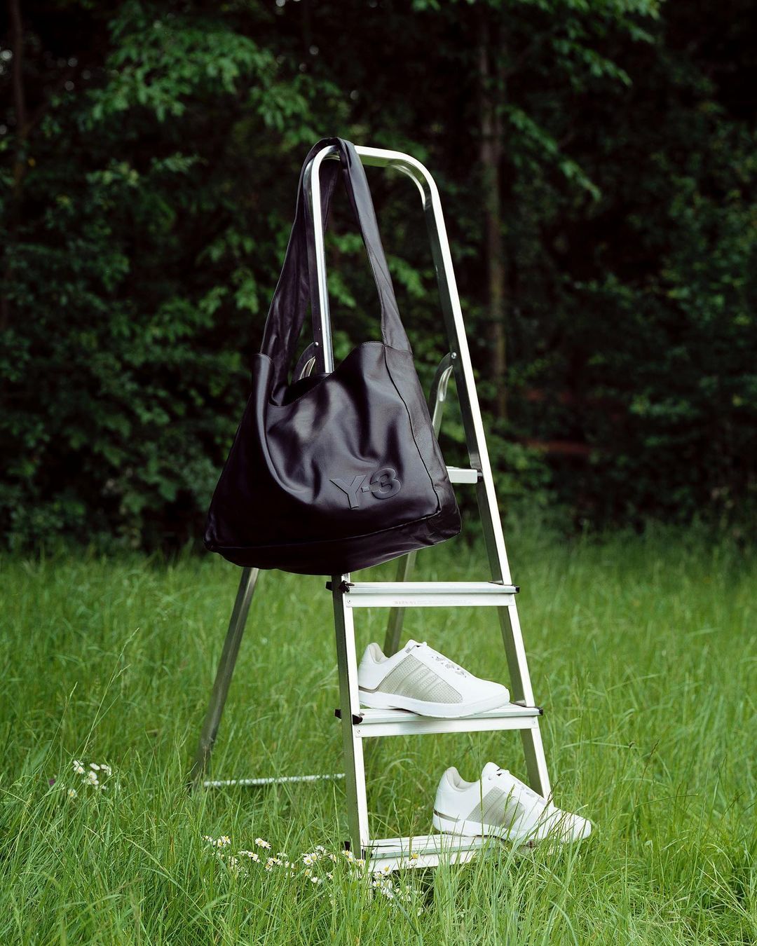 Outdoor fashion shot featuring a black shoulder bag hanging on a metal ladder with white Adidas sneakers in a grassy field.