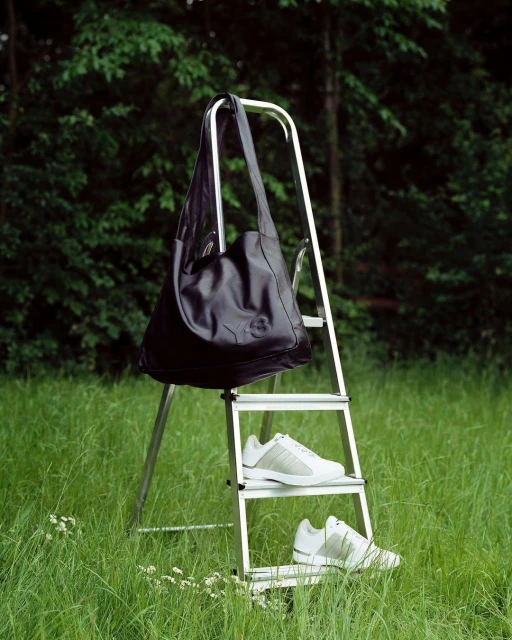 Outdoor fashion shot featuring a black shoulder bag hanging on a metal ladder with white Adidas sneakers in a grassy field.