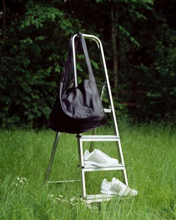Outdoor fashion shot featuring a black shoulder bag hanging on a metal ladder with white Adidas sneakers in a grassy field.