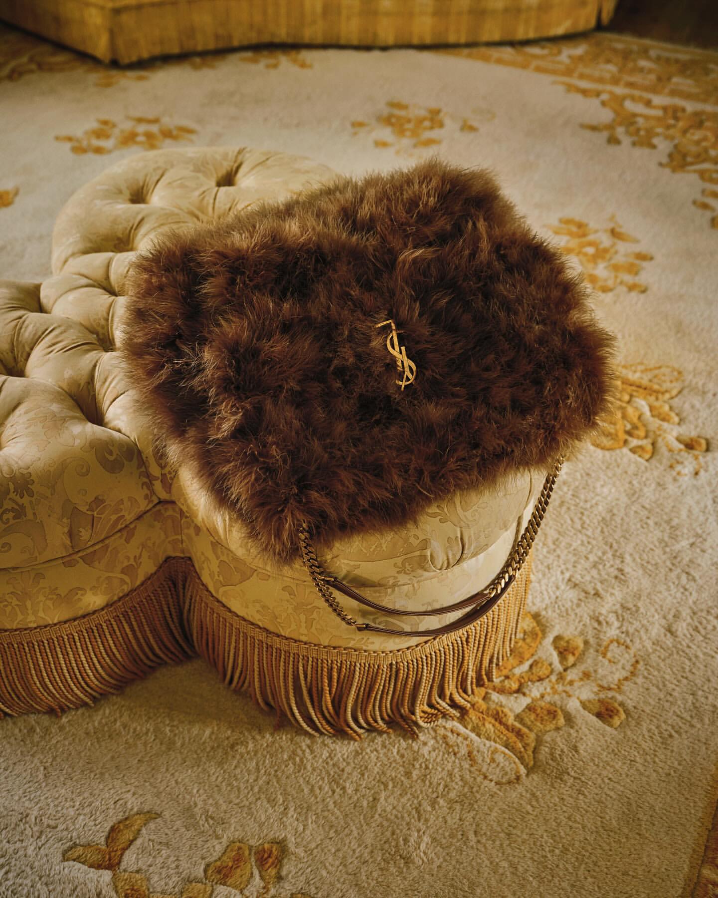 A fluffy brown faux fur handbag with a gold YSL logo rests on an ornate upholstered stool over a patterned carpet.