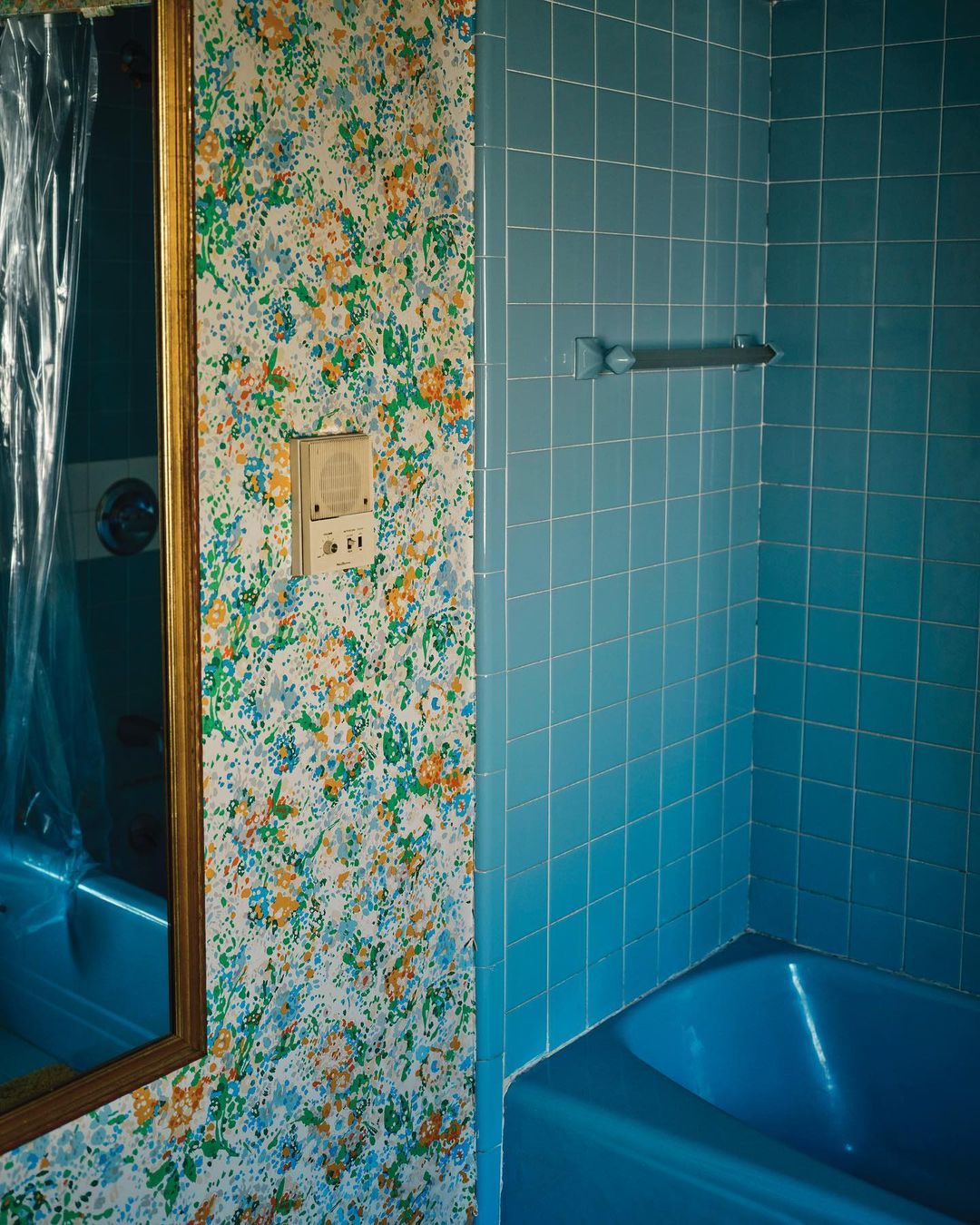 Retro bathroom interior featuring blue tile, floral wallpaper, mirror reflecting a bathtub, and vintage fixtures, evoking a sense of nostalgia.