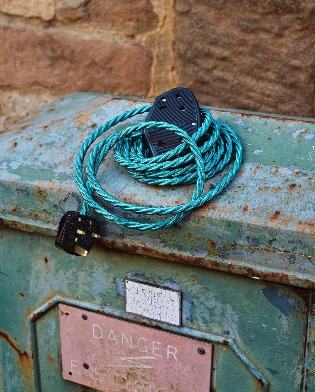 Outdoor shot of a teal electric cable coiled atop a weathered metal box with a 'Danger' sign, set against a rustic brick wall.