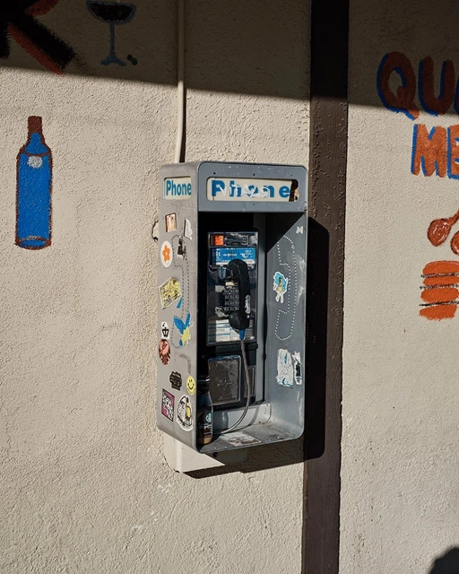 Outdoor photo of a vintage public payphone with stickers attached to the side, mounted on a textured, light-colored building wall.