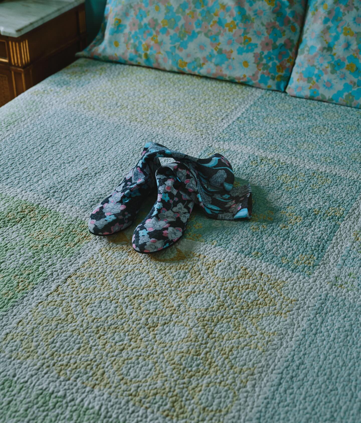 Pair of patterned ankle boots resting on a textured green quilted bedspread with floral pillows