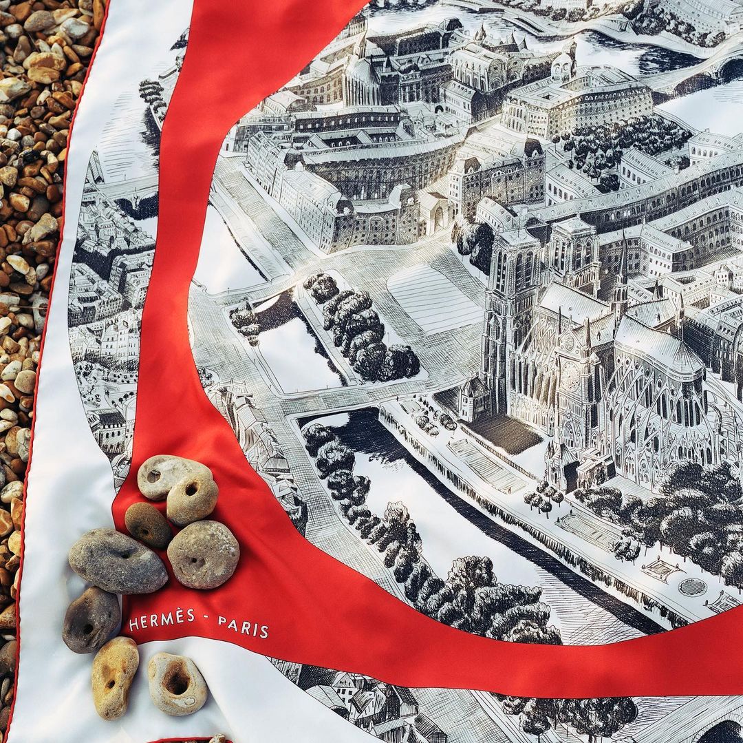 Overhead shot of a silk scarf with an illustrated cityscape design and river, accented with smooth stones on a sandy surface.