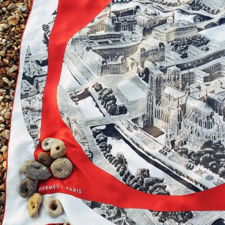 Overhead shot of a silk scarf with an illustrated cityscape design and river, accented with smooth stones on a sandy surface.