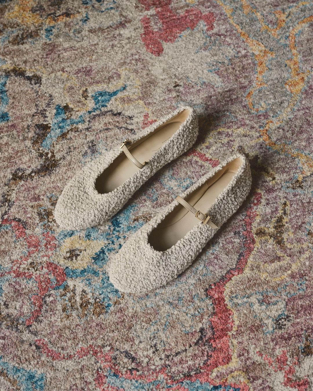 Mary Jane style flats, in a pale cream color, are displayed on a multicolored decorative rug in an overhead shot. Product photography.