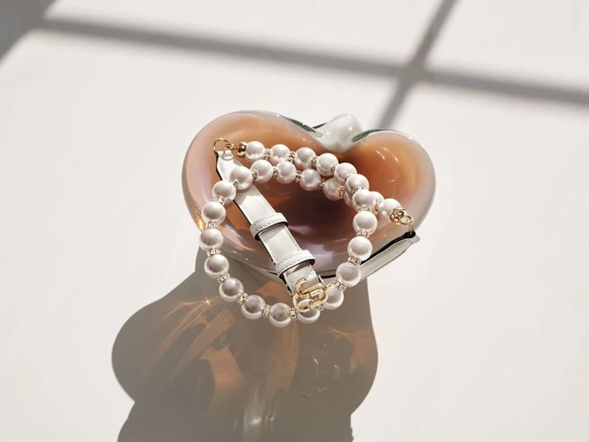 Elegant handbag with pearl strap in an art glass dish, casting shadows on a white surface, showcasing luxury fashion and accessories.