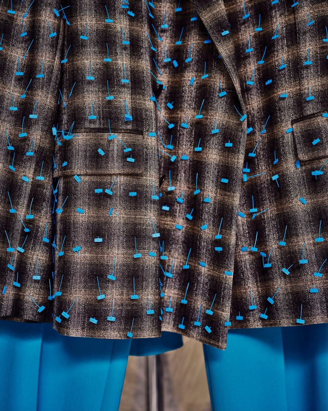 Close-up of a brown plaid jacket with unique turquoise embellishments and matching turquoise leggings, showcasing fashion design.