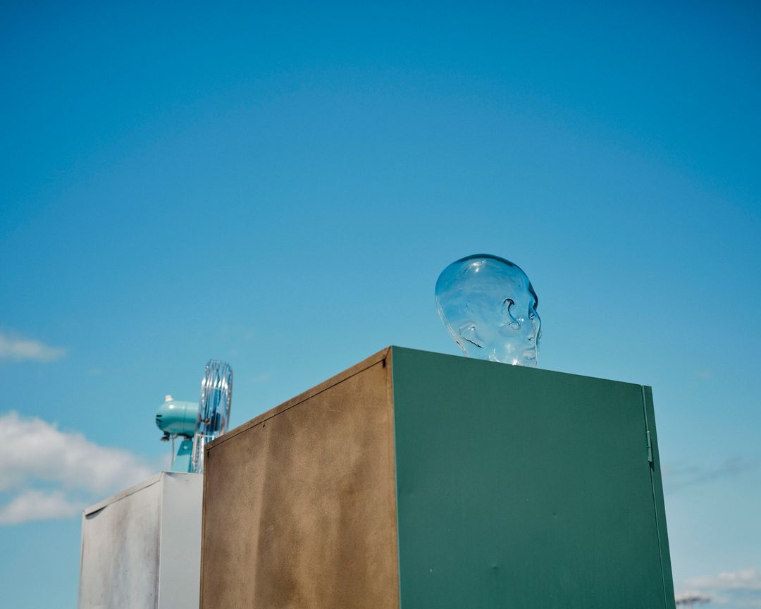 Surreal scene with a transparent skull on a blue block, with a vintage fan and other blocks against a bright blue sky backdrop.
