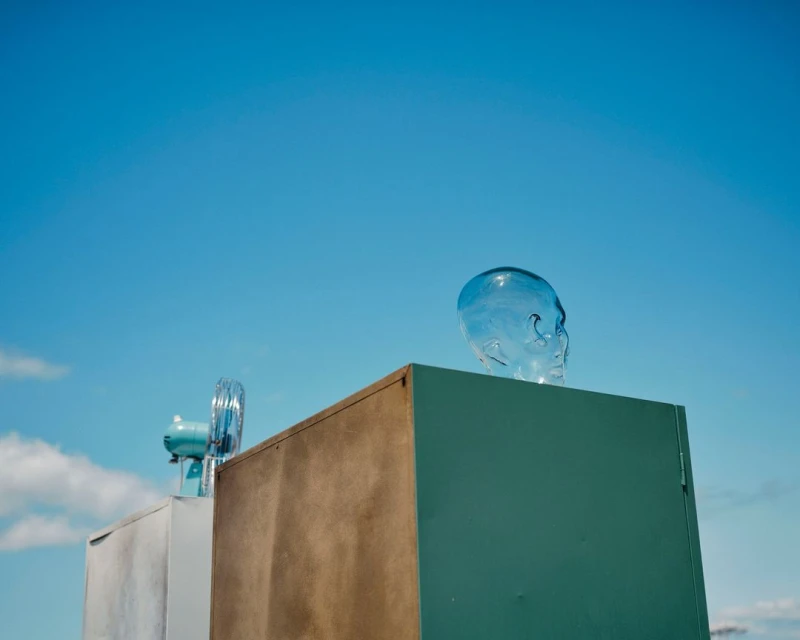 Surreal scene with a transparent skull on a blue block, with a vintage fan and other blocks against a bright blue sky backdrop.
