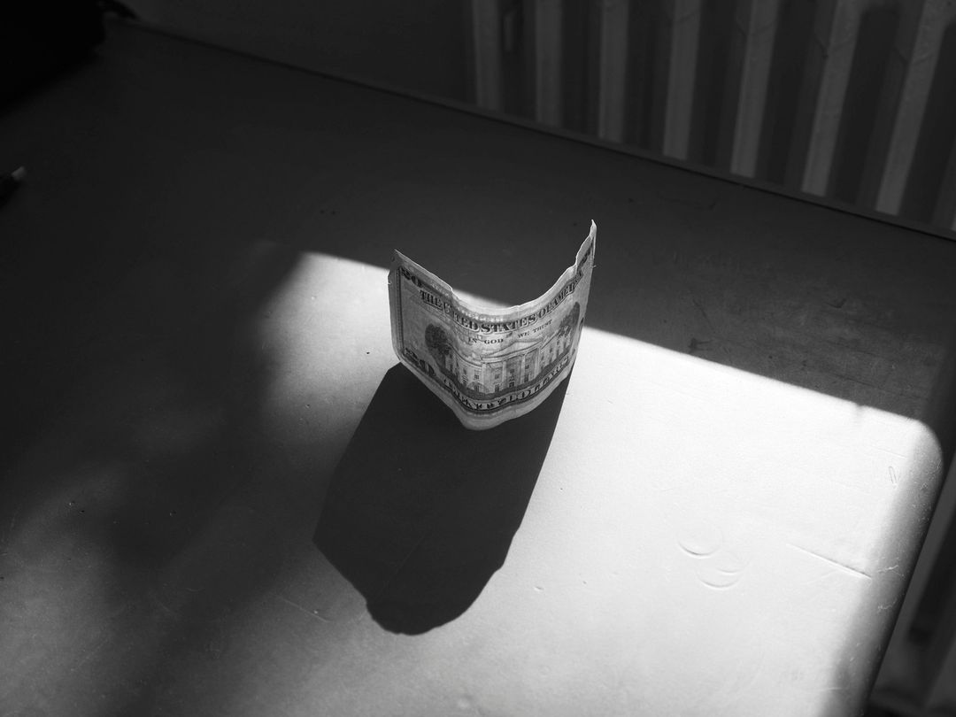 Monochromatic photo of a slightly crumpled U.S. five-dollar bill on a table, casting a distinct shadow in a room with white-striped background.