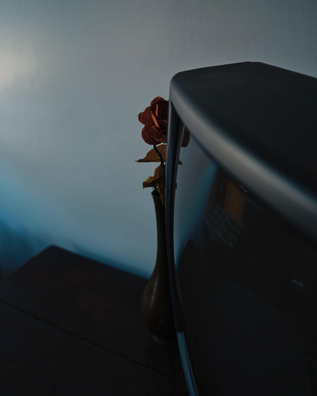 A vintage television sits on a dark wooden table next to an amber vase with a dried rose, set against a blue-grey wall backdrop.