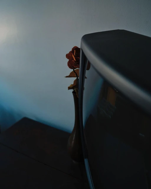 A vintage television sits on a dark wooden table next to an amber vase with a dried rose, set against a blue-grey wall backdrop.