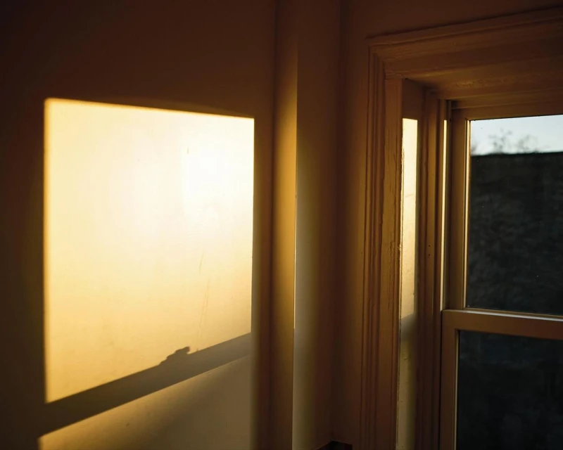 Sunlight creates a bright square on an interior wall next to a window with exterior view. Warm light and shadow create contrast.