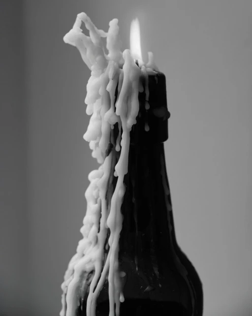 Monochromatic image of a dark glass bottle with melted white wax dripping down its sides and a candle flame at the top.