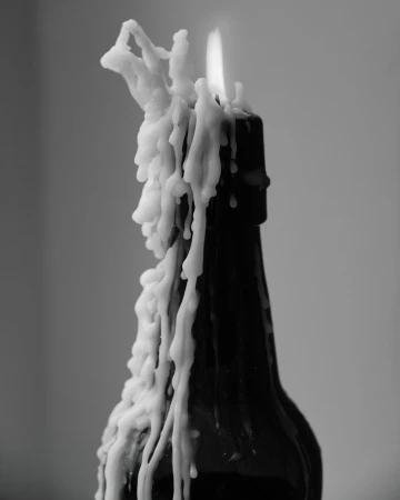 Monochromatic image of a dark glass bottle with melted white wax dripping down its sides and a candle flame at the top.
