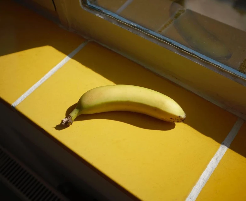 A ripe banana rests on a bright yellow tiled surface near a window, bathed in strong sunlight and casting a distinct shadow.