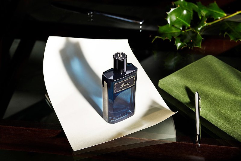 Close-up studio shot of a dark blue Brioni fragrance bottle on white paper with green book and holly, showcasing sophisticated scent.