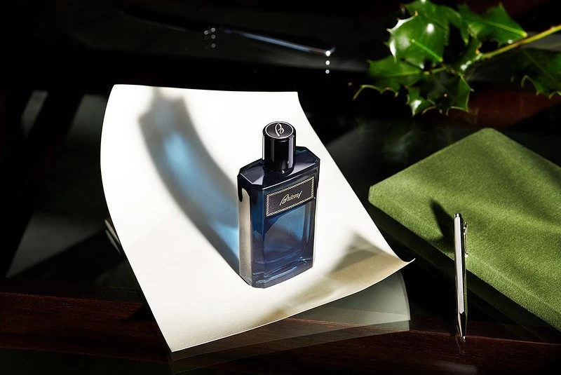 Close-up studio shot of a dark blue Brioni fragrance bottle on white paper with green book and holly, showcasing sophisticated scent.