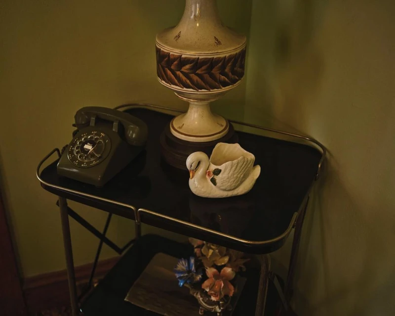 Still life of a vintage rotary telephone, lamp, and swan planter on a metal cart against a muted green wall indoors.