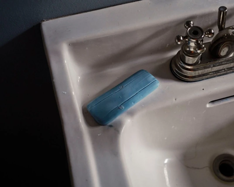 Close-up of a blue bar of soap on a white porcelain sink with a chrome faucet. Dark blue wall is partially visible.