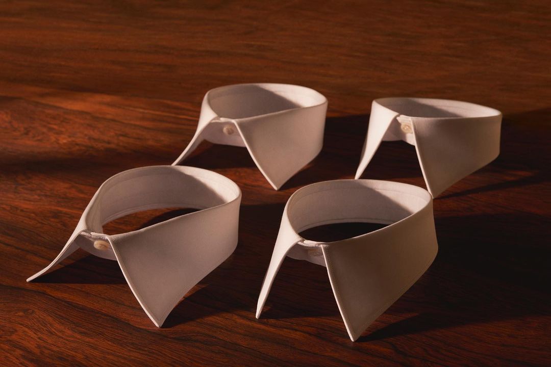 Four white dress shirt collars are neatly arranged on a polished wood surface, evenly lit to highlight their formal design.