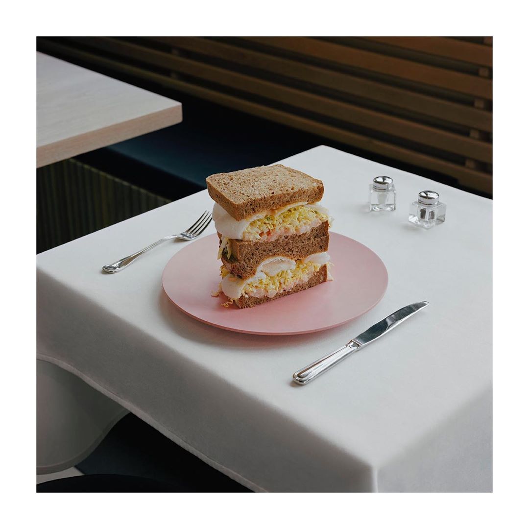 A meticulously plated triple-layer sandwich on a pink plate, silverware and shakers on a white table in a modern dining room.