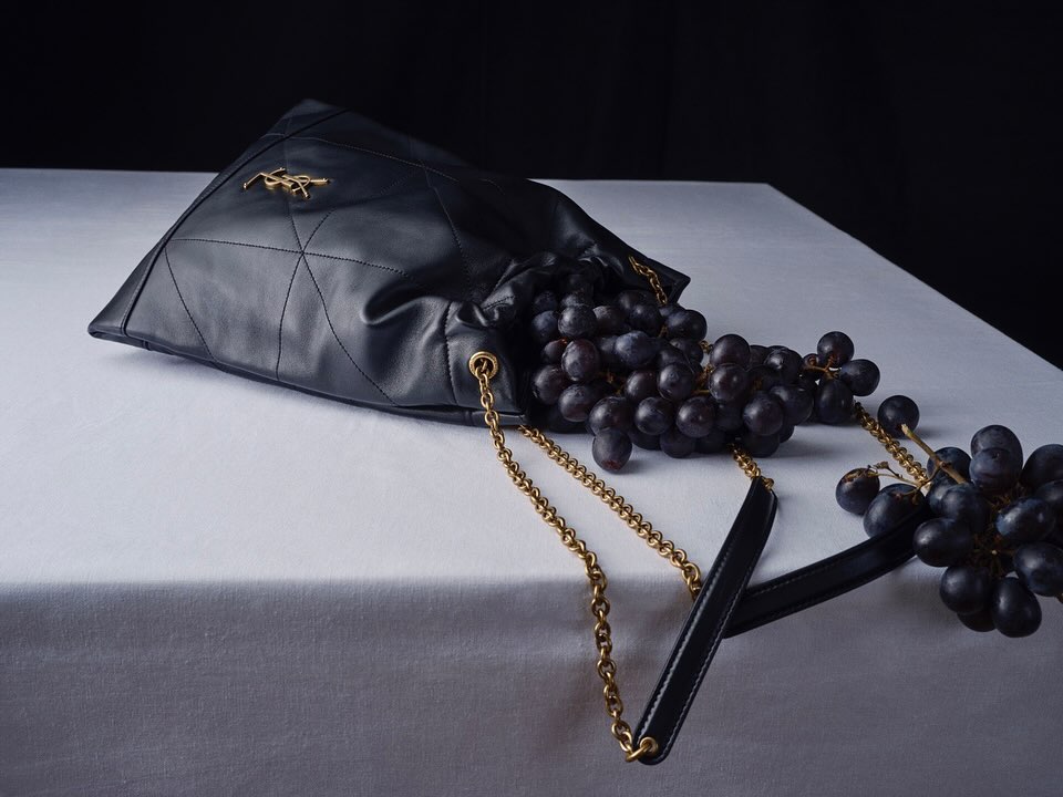 Still life of a black leather handbag overflowing with grapes on a white surface against a dark background in a studio setting.