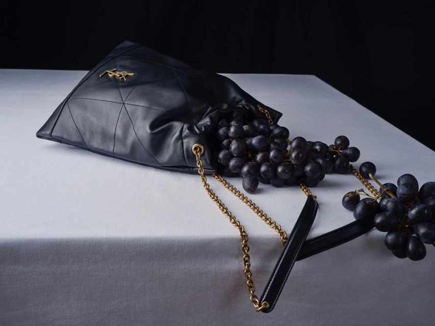 Still life of a black leather handbag overflowing with grapes on a white surface against a dark background in a studio setting.