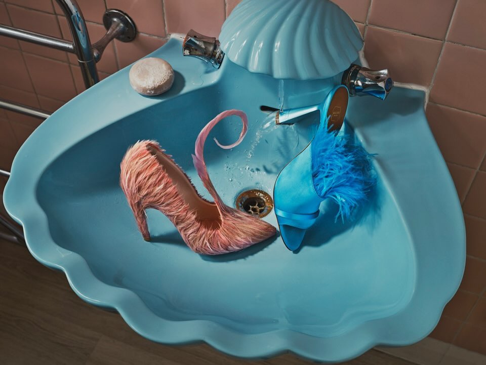 Stylish pink and blue high-heeled shoes in a blue seashell-shaped bathroom sink under a running faucet with pink tile background.