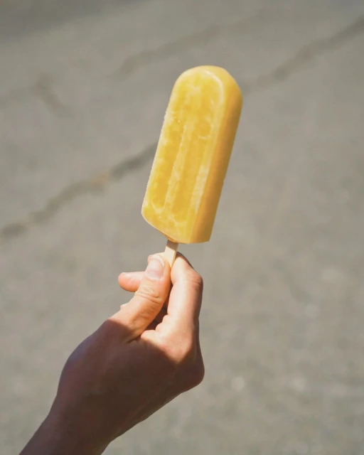 A hand holding a yellow popsicle in bright sunlight against a gray concrete background, showcasing summer refreshment and a simple pleasure.